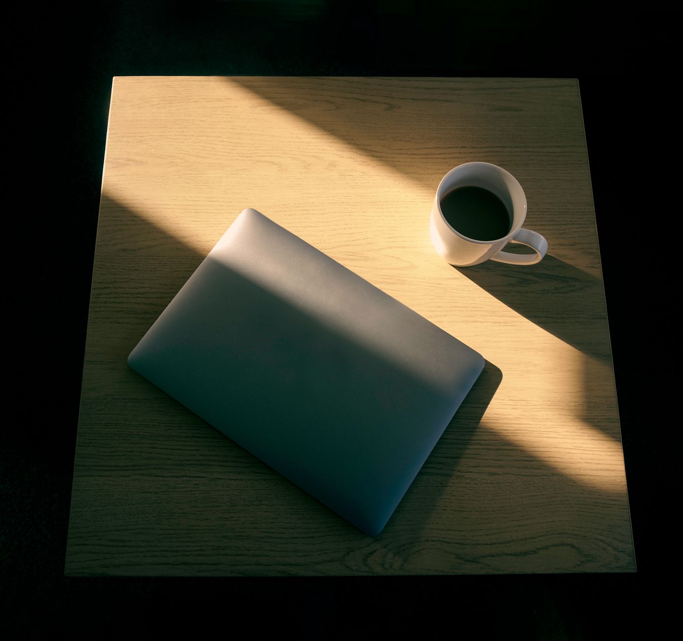Ordinateur portable et tasse de café sur une table en bois, mise en scène minimaliste avec jeu de lumière.